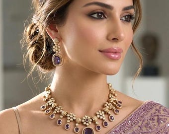 May include: A gold-toned necklace and earring set with purple gemstone accents. The necklace features a large central stone and smaller dangling stones. The earrings have a similar design. The jewelry is worn with a light-colored top and a purple patterned shawl.