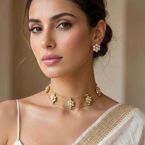 May include: A woman wearing a gold necklace and matching earrings. The necklace is a double-strand choker with flower-shaped pendants. The earrings are also flower-shaped studs. The woman is wearing a white dress.
