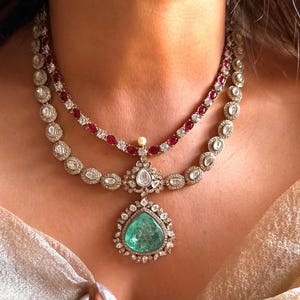 Puede incluir: Un collar de dos hilos con un gran colgante de esmeralda en forma de pera. El collar tiene un engaste de oro con piedras preciosas claras y rojas. El colgante está rodeado por un halo de pequeñas piedras claras.