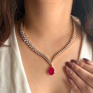 May include: A gold-toned necklace with a sparkling design. The necklace features a series of small, clear stones and a large, teardrop-shaped, red gemstone pendant. The necklace is worn against a white top.