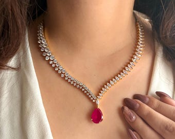 Puede incluir: Un collar dorado con un diseño brillante. El collar presenta un diseño en forma de V con piedras claras y brillantes y un gran colgante rojo en forma de lágrima. El collar lo lleva una persona que lleva una blusa blanca.