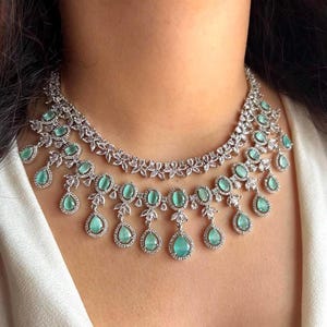 Op de afbeelding: Zilverkleurige ketting met twee strengen, versierd met lichtgroene, druppelvormige edelstenen. De ketting heeft een bloemenontwerp en is versierd met kleinere, heldere stenen. Het ontwerp is delicaat en elegant.