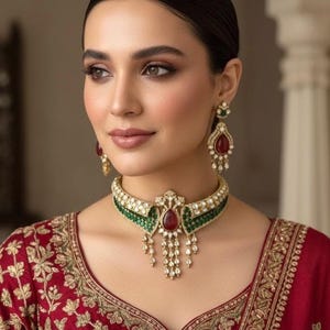 May include: A woman wearing a gold-toned necklace and matching earrings. The necklace features a green and white band with a large red gemstone pendant and dangling accents. The earrings have a similar design with red gemstones. The woman is wearing a red top with gold embroidery.