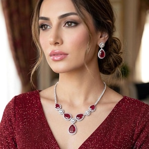 Può includere: Un abito rosso scintillante è abbinato a un set di collana e orecchini di rubini e diamanti abbinati. La collana presenta un pendente a goccia e una serie di pietre color rubino circondate da diamanti. Gli orecchini hanno un design simile.