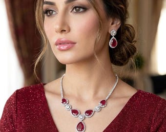 Puede incluir: Un vestido rojo brillante se combina con un conjunto de collar y pendientes de rubíes y diamantes a juego. El collar presenta un colgante en forma de lágrima y una serie de piedras de color rubí rodeadas de diamantes. Los pendientes tienen un diseño similar.