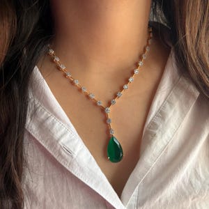 May include: A gold necklace with a teardrop-shaped green gemstone pendant. The necklace features a delicate chain with small, sparkling accents. The pendant is a vibrant green color, and the necklace is worn against a white shirt.