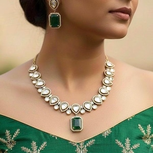 May include: A gold-toned necklace and earring set featuring emerald green and clear gemstones. The necklace has a pendant with a rectangular emerald, and the earrings have a similar design. The jewelry is set against a green top with gold embroidery.