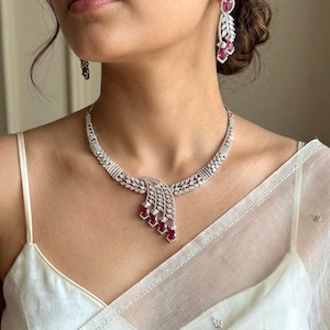 Può includere: Una collana d'argento e un set di orecchini abbinati con pietre preziose chiare e rosse. La collana presenta un design a cascata con un gruppo centrale di pietre rosse. Gli orecchini hanno un design simile, con gemme pendenti.