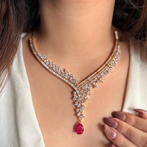 以下が含まれることがあります： 赤い宝石がついた涙型のペンダントが付いた金のネックレス。ネックレスは透明なキュービックジルコニアで飾られています。