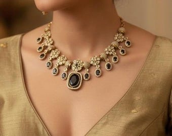 May include: A gold-toned necklace and matching earrings set. The necklace features a central black oval stone, surrounded by smaller black stones and floral accents. The earrings have a similar design. The jewelry is worn over a gold-colored top.