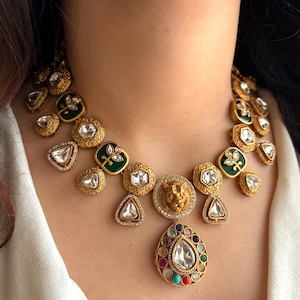 May include: A gold necklace with a lion's head pendant and various gemstone accents. The necklace features a teardrop-shaped pendant with a large central stone and smaller colourful stones around it. The necklace also has green enamel and clear stone details.