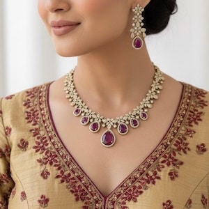 May include: A gold-toned necklace and earring set with ruby-colored teardrop-shaped stones and clear accent stones. The necklace is a floral design. The earrings are a matching floral design. The woman is wearing a gold-colored top with burgundy embroidery.