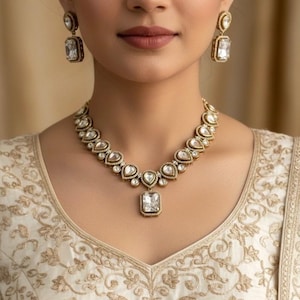 May include: A gold-toned necklace and matching earrings set. The necklace features a series of heart-shaped and rectangular clear stones. The earrings have a rectangular stone. The jewelry is worn over a cream-colored embroidered top.