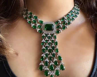 May include: A dazzling necklace featuring a large, square emerald-cut green gemstone at the center, surrounded by smaller emerald-cut and pear-shaped green gemstones. The necklace has a silver-colored setting and a cascading design.