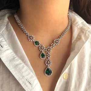 May include: A stunning necklace with clear, sparkling stones and vivid green, teardrop-shaped gems. The necklace is set against a white shirt, accentuating its detailed design and elegant appearance. A statement piece of jewellery.