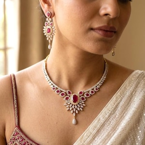 May include: A necklace and matching earring set featuring oval and teardrop-shaped red gemstones surrounded by small white stones set in a metallic frame. The necklace has a central pendant design with a dangling stone, and the earrings are drop style.