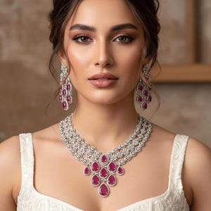 May include: A woman wearing a white dress and a sparkling necklace and earring set. The jewelry features clear stones and teardrop-shaped ruby-colored gems. The necklace has multiple layers, and the earrings dangle.