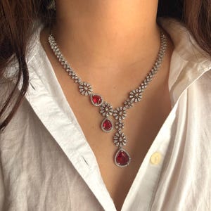 May include: A silver necklace with a teardrop-shaped red gemstone pendant. The necklace is adorned with small, clear gemstones arranged in a floral pattern.