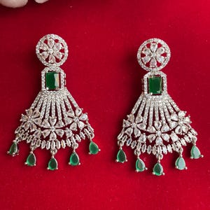 May include: A pair of silver earrings with a green stone and a floral design. The earrings have a dangling design with multiple layers of small stones.