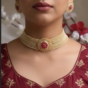 Può includere: Una collana di perle e un set di orecchini abbinati. La collana è un girocollo a più fili con una gemma rossa centrale. Gli orecchini sono rotondi con una pietra rossa al centro e dettagli dorati. La donna indossa un top bordeaux con ricami dorati.