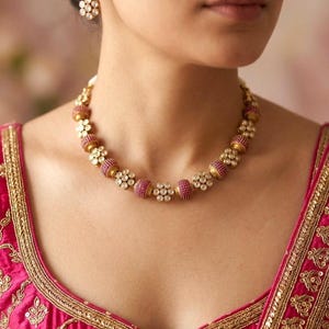 Könnte beinhalten: Ein goldfarbenes Halsketten- und Ohrring-Set. Die Halskette hat abwechselnd rosa und goldfarbene Perlen mit floralen Akzenten. Die Ohrringe haben ein ähnliches Design. Das Model trägt ein rosa besticktes Oberteil.