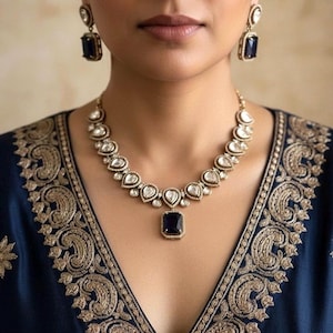 May include: A gold-toned necklace and matching earrings set with clear and dark blue gemstones. The necklace features a central rectangular dark blue stone pendant. The earrings have a similar design. The model is wearing a dark blue top with gold embroidery.
