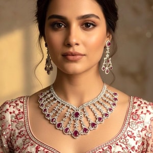 May include: A woman wearing a diamond necklace and matching earrings with ruby-colored gemstones. The necklace has a detailed, openwork design. The woman is wearing a white top with red floral embroidery.