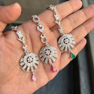 May include: Three silver and white jeweled teardrop pendants with a circular design and a small colored gemstone hanging from the bottom. The gemstones are pink, red, and green.