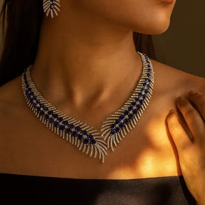 Può includere: Un'affascinante collana e orecchini coordinati. I gioielli presentano un design a forma di piuma con file alternate di pietre preziose blu e pietre bianche scintillanti. La collana incornicia graziosamente il décolleté.