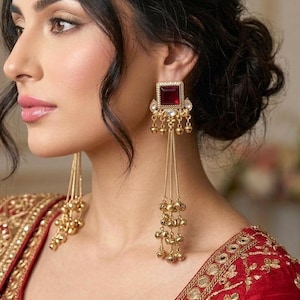 May include: Gold-toned earrings with a square, deep red gemstone center and dangling chains of small gold beads. The earrings are part of a set with a red and gold embroidered garment.