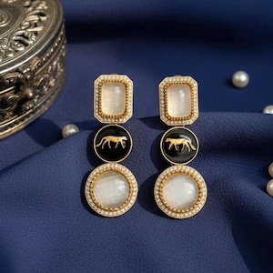 May include: Gold-toned drop earrings with a three-tiered design. The top tier features a rectangular, clear stone surrounded by small crystals. The middle tier has a black circle with a gold animal silhouette. The bottom tier has a round, clear stone.