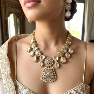 May include: A gold-toned necklace and matching drop earrings featuring translucent, oval-shaped stones set in ornate metal frames. The necklace includes a large, trapezoidal central pendant with intricate floral detailing and smaller stone accents.