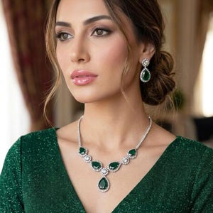 Può includere: Una donna indossa un abito verde e un set di gioielli abbinato con smeraldi e diamanti. La collana presenta smeraldi a goccia circondati da diamanti. Anche gli orecchini hanno smeraldi e diamanti. La donna ha i capelli castani e guarda di lato.