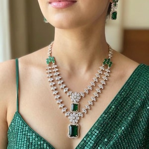 Puede incluir: Un collar de dos capas con piedras rectangulares de color verde esmeralda y piedras blancas transparentes de corte marquesa dispuestas en forma de hoja, con pendientes a juego. Se lleva con una prenda verde sin mangas con lentejuelas.