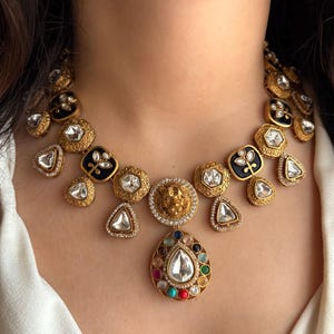 May include: A gold-toned statement necklace featuring a central teardrop pendant with a large, clear stone surrounded by colorful gems. The necklace includes various shapes, including black enamel squares and clear, faceted stones.