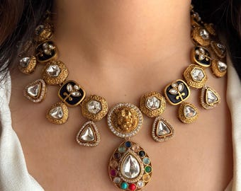 May include: A gold-toned statement necklace featuring a central teardrop pendant with a large, clear stone surrounded by colorful gems. The necklace includes various shapes, including black enamel squares and clear, faceted stones.