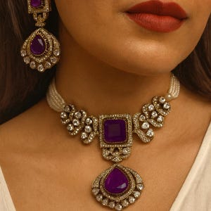 以下が含まれることがあります： ネックレスとイヤリングが特徴のジュエリーセットのクローズアップ。セットには、正方形の紫色の宝石ペンダントと涙の形をしたペンダントが付いたネックレスが含まれています。イヤリングには、お揃いの正方形の紫色の宝石が付いています。ジュエリーはゴールドトーンです。