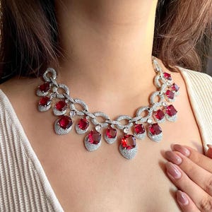 Puede incluir: Un collar deslumbrante con piedras rojas cuadradas, intercaladas con cristales transparentes más pequeños. El metal plateado resalta las vibrantes piedras rojas. El diseño es elegante y llamativo, ideal para ocasiones especiales.