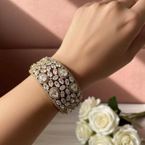 May include: A gold-toned bracelet with a dazzling array of clear, sparkling gemstones. The bracelet features a wide band design, with the gemstones set closely together. The overall effect is one of luxury and elegance, perfect for special occasions.
