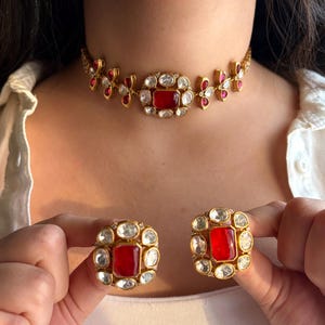 May include: Gold-toned jewelry set featuring a choker necklace and matching earrings. The necklace and earrings have a central red gemstone surrounded by clear stones. The necklace has a floral design with red accents.