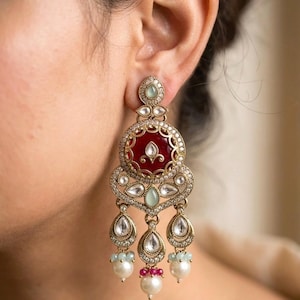 May include: Close-up of an ornate gold earring with a red center and clear and pale green accents. The earring has a teardrop design with dangling pearls and small pink and blue beads. The earring is worn on a person's ear.