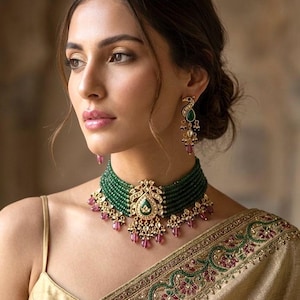 May include: A woman wearing a gold-colored sari and a green beaded choker necklace with a central pendant and matching earrings. The necklace has pink accents and the earrings feature a peacock design.