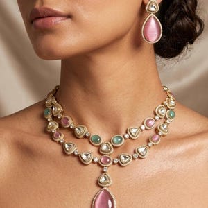 Puede incluir: Un conjunto de collar y pendientes dorados. El collar presenta piedras preciosas rosas, verdes y transparentes. Los pendientes tienen una piedra preciosa rosa en forma de lágrima. La joyería está sobre un fondo neutro.