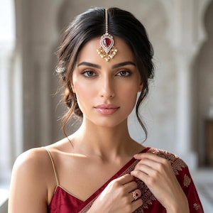 May include: A gold-toned headpiece with a central red gemstone and surrounding white stones and pearls. The headpiece is worn on the forehead. The image also shows a red ring with a matching gemstone and a red garment with gold embroidery.