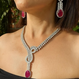 Può includere: Un set di gioielli abbagliante con collana e orecchini abbinati. La collana e gli orecchini sono argentati e adornati con scintillanti pietre trasparenti e grandi pietre preziose rosso rubino di forma ovale. Gli orecchini sono lunghi e pendenti, mentre la collana ha un design unico.