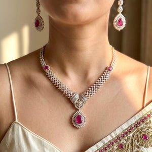 May include: A necklace and matching drop earrings set featuring teardrop-shaped pink stones surrounded by small clear crystals. The necklace has a central teardrop pendant and a band of intricate crystal settings, while the earrings are long.
