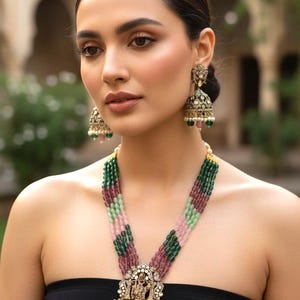 Könnte beinhalten: Eine mehrsträngige Halskette mit einem großen Anhänger und passenden Ohrringen. Die Halskette hat abwechselnde Stränge aus grünen, rosa und burgunderfarbenen Perlen. Der Anhänger hat ein detailliertes Golddesign mit kleinen weißen und farbigen Akzenten. Die Ohrringe haben ein ähnliches Design.