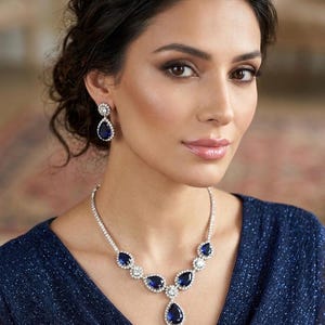 Può includere: Un set di gioielli abbagliante con collana e orecchini abbinati. La collana presenta una serie di pietre a forma di goccia blu scuro, intervallate da pietre più piccole e trasparenti, incastonate in un metallo argentato. Gli orecchini riprendono il design della collana.