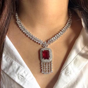 Può includere: Una collana d'argento con un grande pendente rettangolare di pietra rossa. Il pendente è circondato da pietre più piccole e trasparenti e ha una frangia di pietre pendenti. La collana è indossata su una camicia bianca.