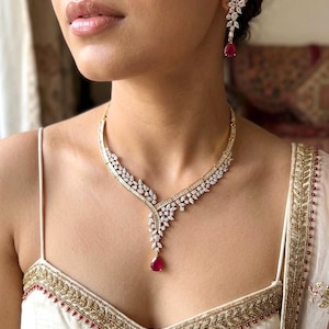 Può includere: Un set composto da collana e orecchini pendenti color oro con pietre trasparenti taglio baguette e marquise disposte a motivo floreale, con una singola pietra rosso rubino a goccia sospesa al centro della collana e su ogni orecchino.